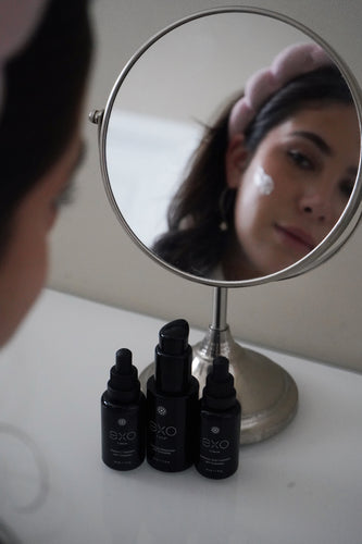 A morning begins in reflection: Exoceuticals® bottles meet the quiet gaze of a woman in the mirror, capturing the rhythm of radiant self-starts.
