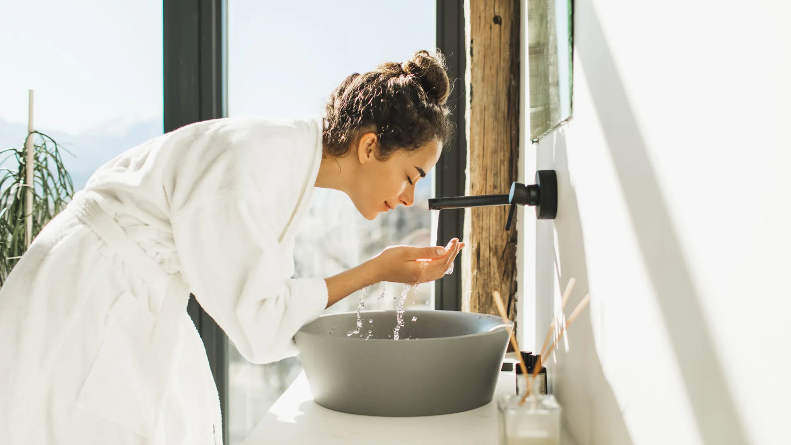 A skinimalist skincare ritual in progress — woman in a robe preparing to cleanse her face at a modern sink, illustrating the “less is more” beauty trend.