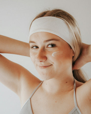 Smiling and sunlit, a young woman applies under-eye cream — a visual emblem of age-aligned skincare awareness and preventative care culture.
