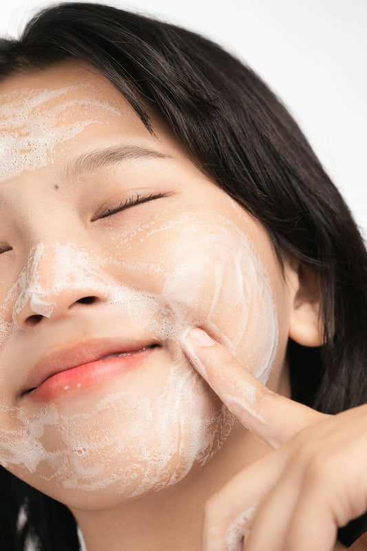 With closed eyes and gentle pressure, a woman massages exfoliant into her skin — a visual cue for timing, touch, and the care behind every cleanse.